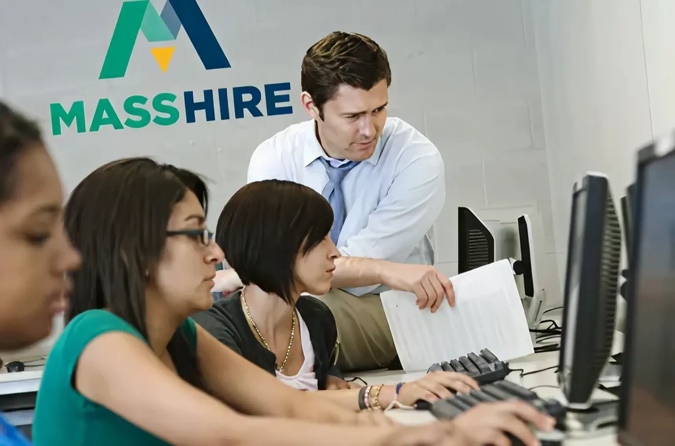 一个专业的工作空间，个人专注于电脑，一名工作人员提供指导，背景是 "MassHire "标志。