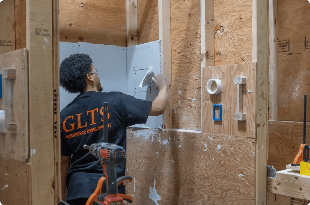 在建筑培训场所，一名身穿 GLTS 衬衫的工人正在给干墙涂抹饰面胶，周围是工具和木板。