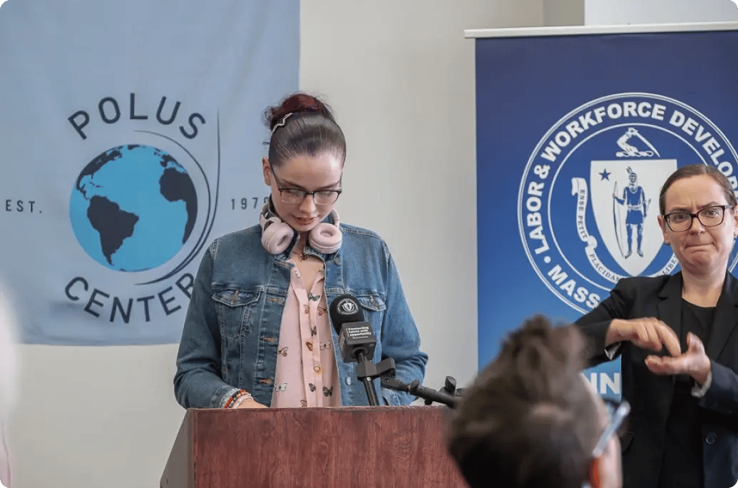 一位演讲者站在讲台上，面前是波卢斯中心的横幅，一旁还有一位手语翻译。