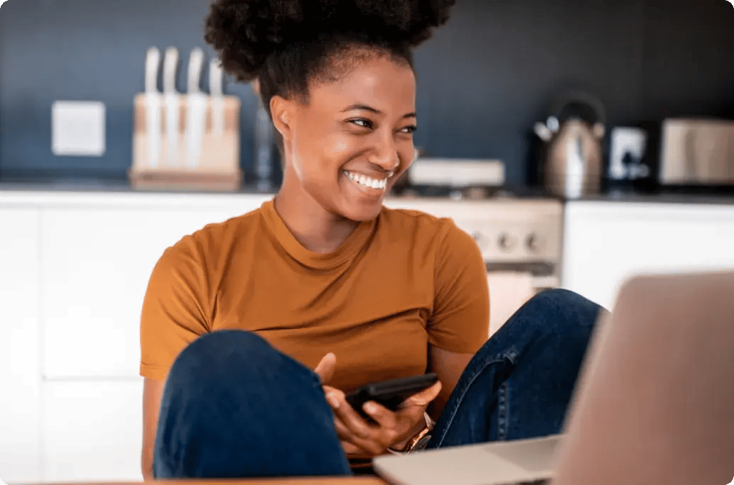 在家中使用笔记本电脑工作的人，在明亮的厨房工作区一边使用手机一边微笑。