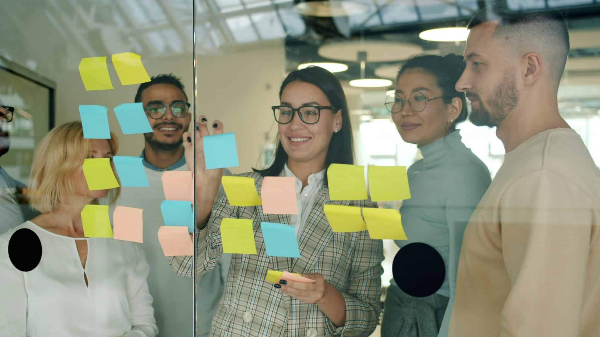 A group of people gathers around a glass wall covered in colorful sticky notes, discussing ideas and collaborating